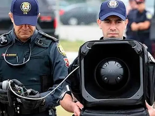 Dos agentes de policía sujetando un LRAD 100X, un altavoz acústico portátil de color negro.